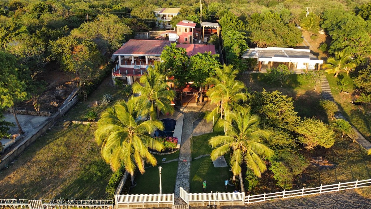 An aerial view reveals a spacious two-level beach house surrounded by lush greenery. The property features a well-kept garden with palm trees, a pathway leading to a pool, and multiple buildings, including a charming outdoor area for gatherings.