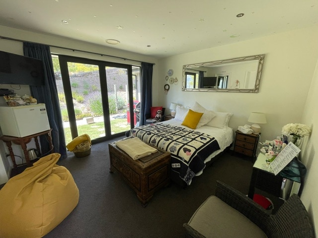 A spacious bedroom features a large bed covered with a patterned blanket, and a mirror reflecting natural light. A cozy chair and a small table are positioned next to a window, and a mini fridge is placed in the corner.
