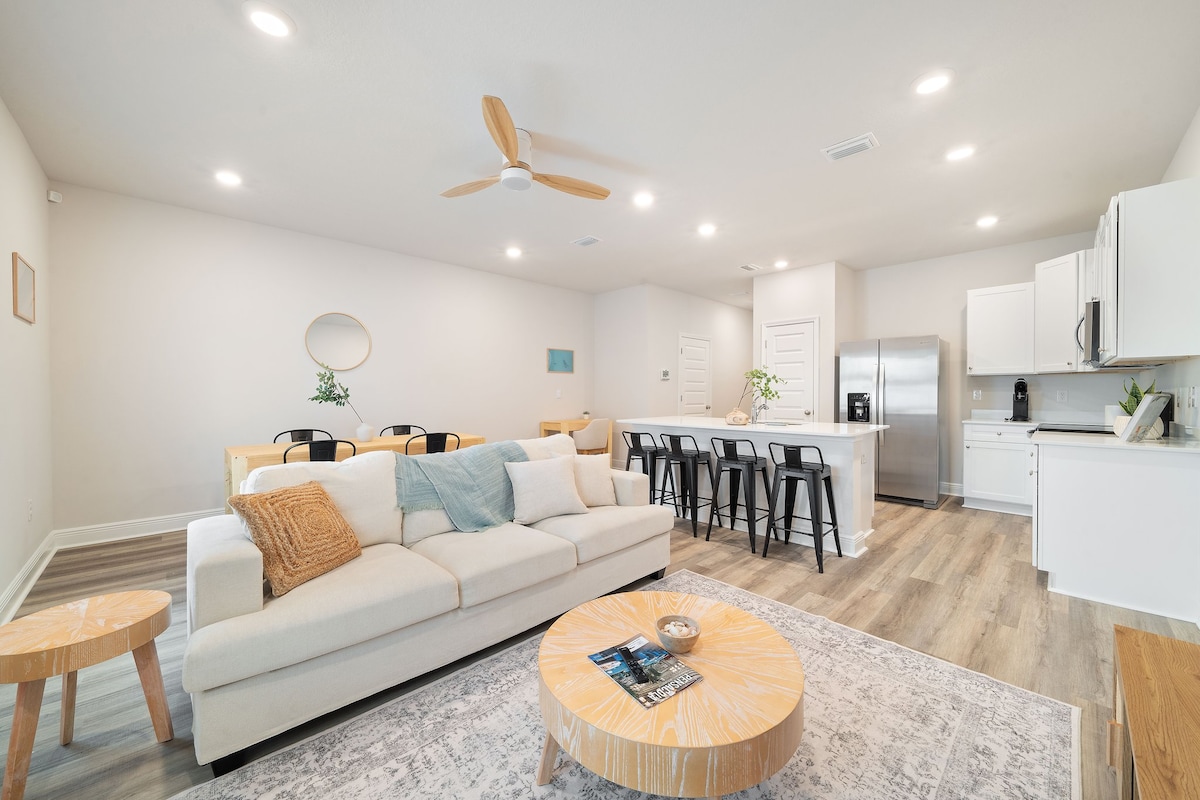 A modern open-concept living space features a light-colored sectional sofa centered on a round coffee table. The kitchen is visible in the background, showcasing sleek cabinetry and a large island with bar seating. Dining chairs are arranged at a table near the window.