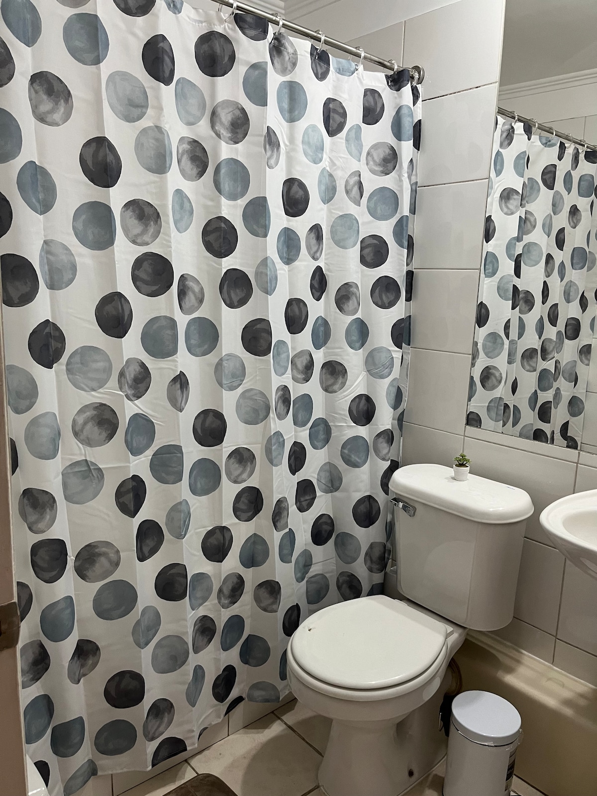 A bathroom is featured with a white tiled design. A shower curtain adorned with varying-sized blue and gray circles provides privacy. A compact toilet and a sink with a mirror are positioned adjacent to the shower area, contributing to the overall functionality of the space.