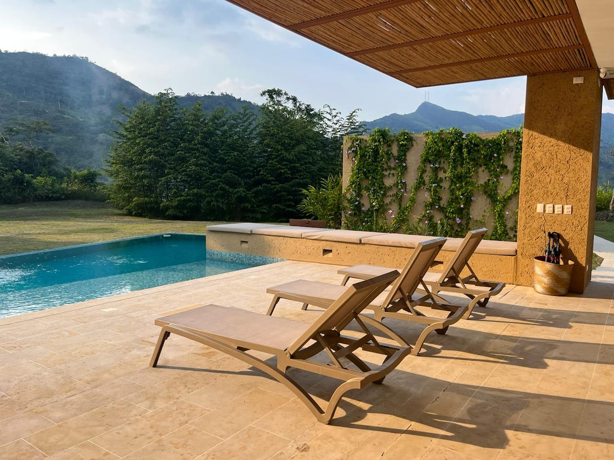 A poolside area is presented with several lounge chairs arranged near a clear blue pool. Lush greenery borders the space, complemented by a backdrop of gentle hills under a clear sky. Natural light enhances the serene atmosphere of this outdoor retreat.