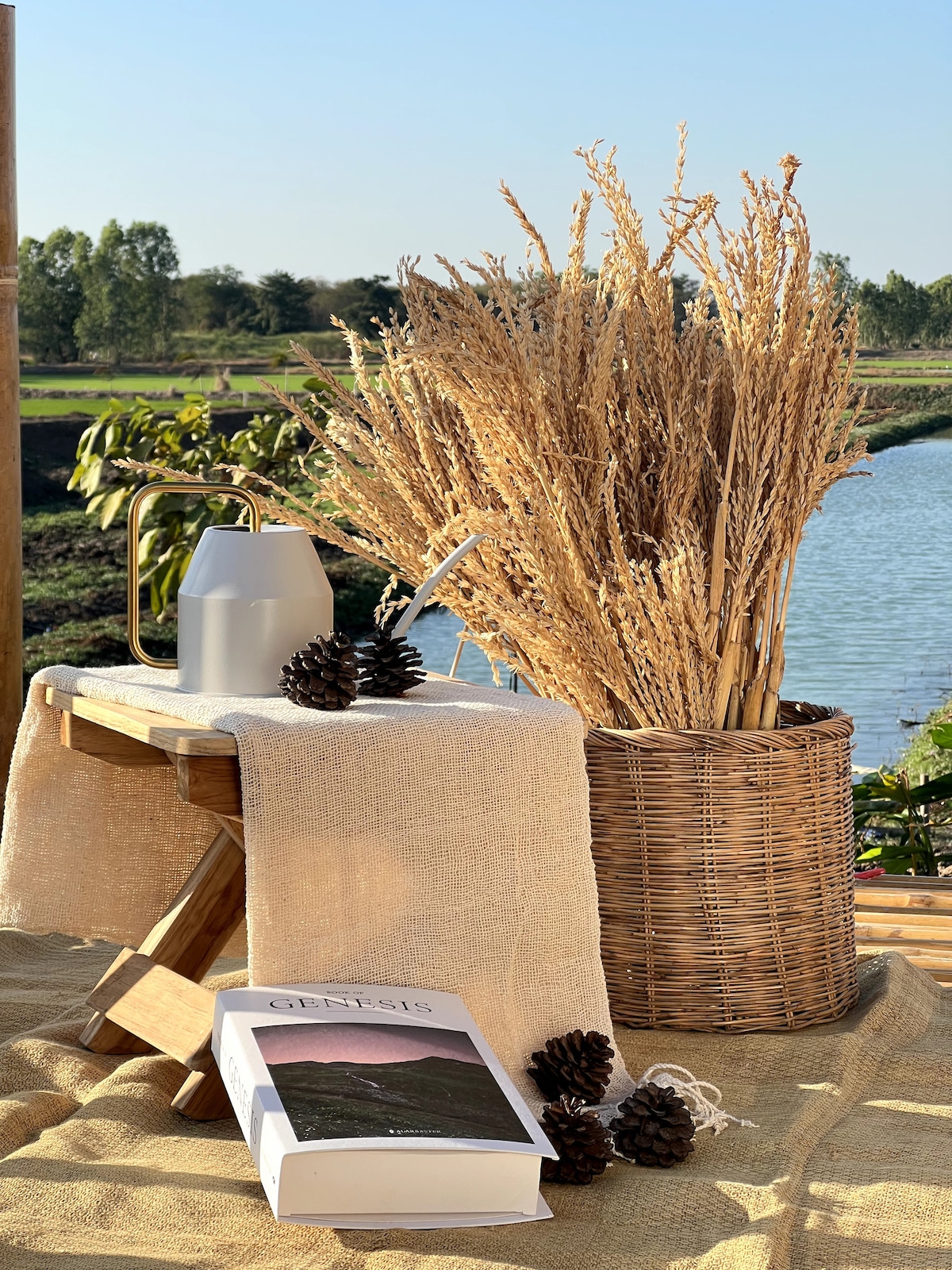 A wooden stool is placed beside a basket filled with dried grass, enhancing the natural ambiance. A metal pitcher sits on the table, accompanied by a textured fabric draped over it. In the background, a serene waterway reflects the surrounding greenery.