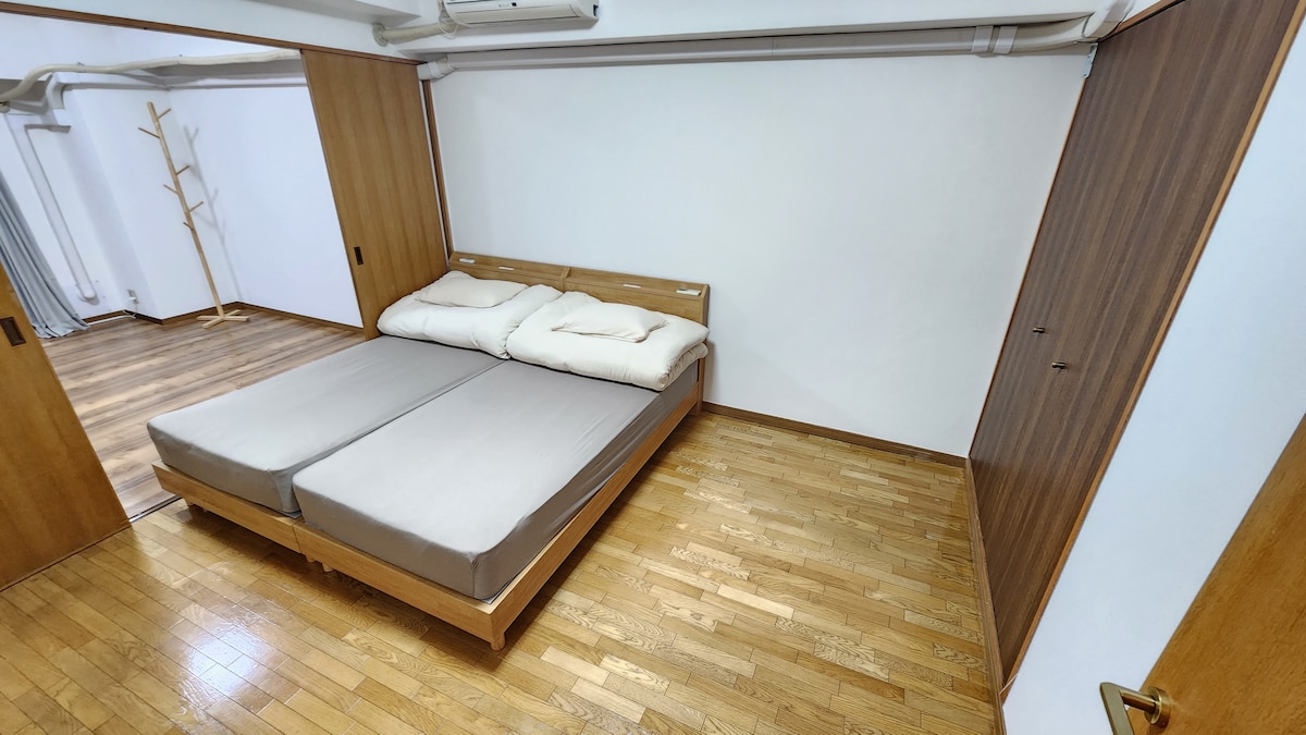 A spacious room is presented with a large, double queen-sized bed featuring light-colored bedding atop a wooden frame. The polished wooden floor complements the minimalistic design, while soft natural light gently fills the area from the entrance.