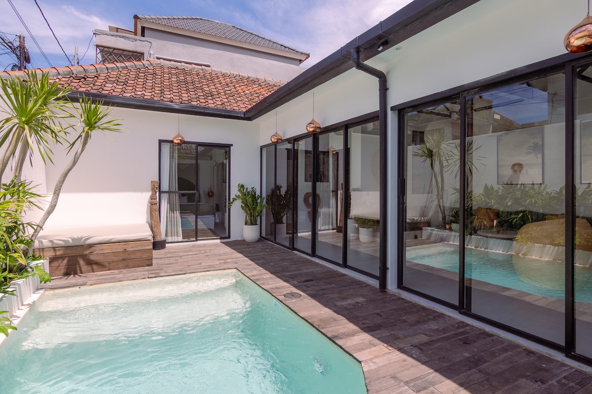 A serene outdoor space features a tranquil pool with clear water, surrounded by wooden decking and tropical plants. Large glass doors reflect bright natural light, seamlessly connecting indoor and outdoor areas. Elegant pendant lights add a touch of sophistication to the atmosphere.