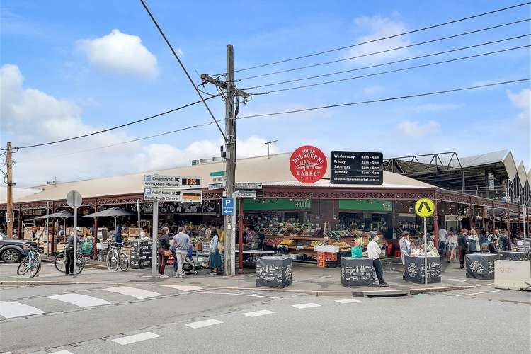 A bustling market scene reveals a large building with a prominent sign displaying ‘South Melbourne Market.’ Stalls brimming with fresh produce and various goods are visible. People are seen shopping and interacting, while bicycles are parked nearby, reflecting the vibrant community atmosphere.