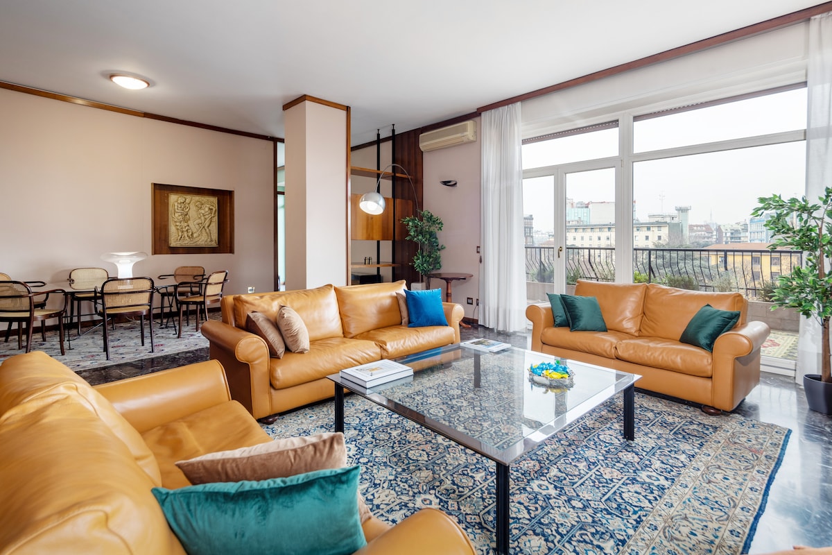 A spacious living area is furnished with three leather sofas and a decorative glass coffee table, surrounded by a richly patterned rug. Large windows provide ample natural light and scenic views of the city. A dining table with chairs is visible in the background.