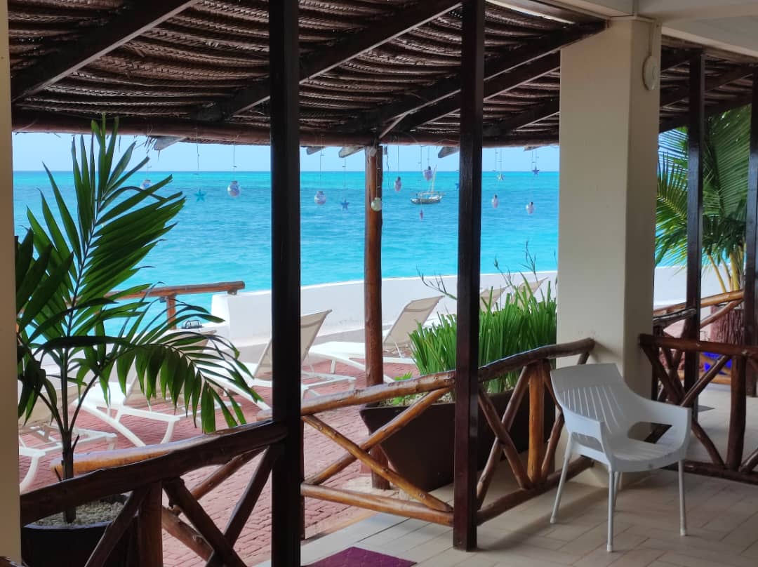 A covered outdoor seating area is visible, featuring a white chair positioned beside lush green plants. The view beyond showcases the blue waters of the Indian Ocean, with a gentle shoreline and a boat anchored in the distance. Baskets hang from the structure's ceiling.