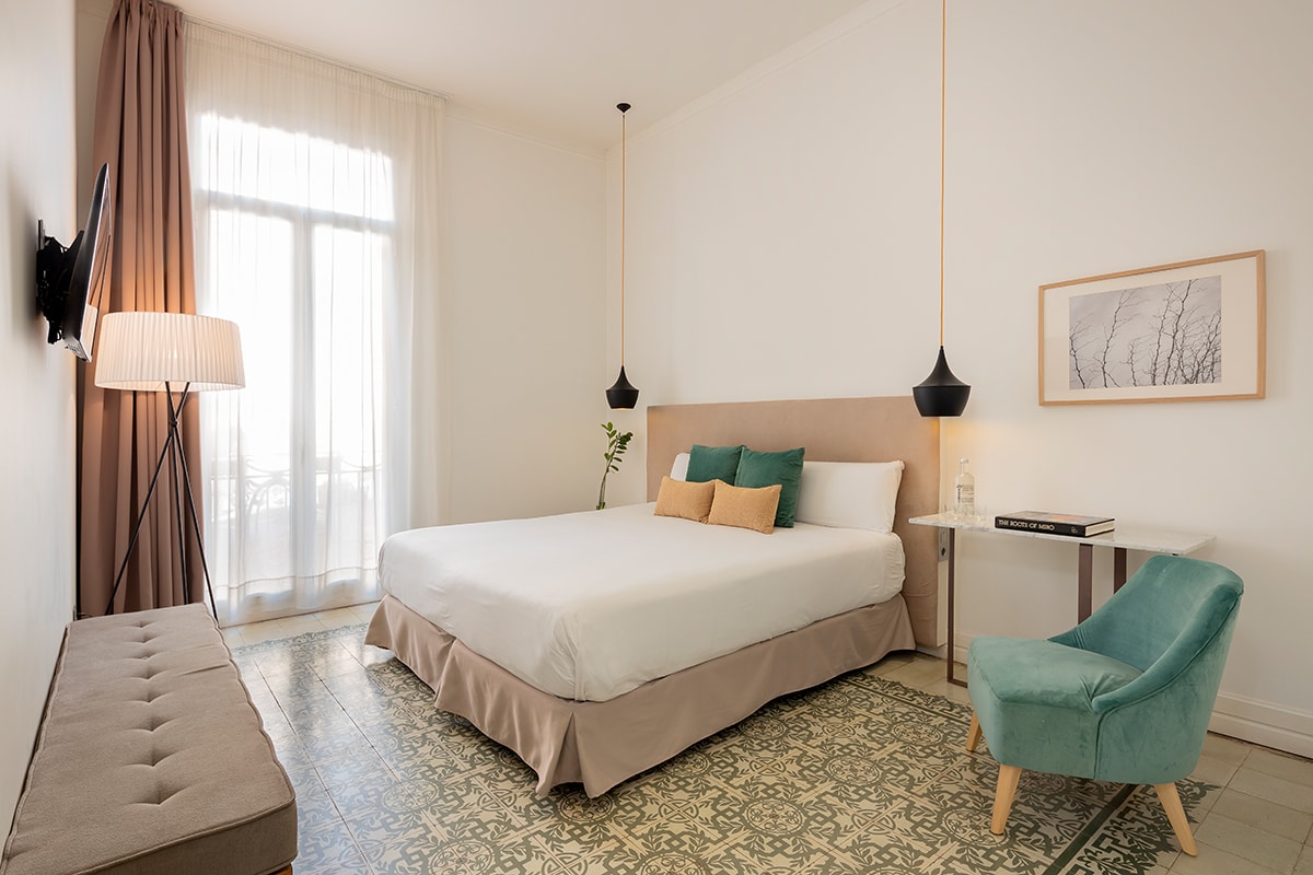 A bright room features a spacious bed with neutral bedding and decorative throw pillows. Soft natural light filters through a large window covered by sheer curtains. A small green chair and a bench are positioned beside a minimalist side table, enhancing the room's calming atmosphere.