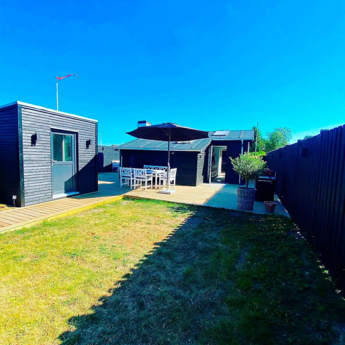 A spacious outdoor area is featured, showcasing a large wooden terrace with a dining table and seating under an umbrella. The surrounding garden is well-kept, with grassy areas and potted plants. Two black wooden structures provide a modern aesthetic against a clear blue sky.