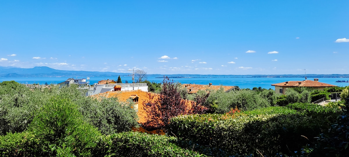 A panoramic view showcases serene blue waters of Lake Garda, bordered by vibrant green trees and lush shrubs. Terracotta rooftops are visible in the foreground, merging harmoniously with the natural landscape. The mountains in the distance complete this tranquil setting under a clear blue sky.