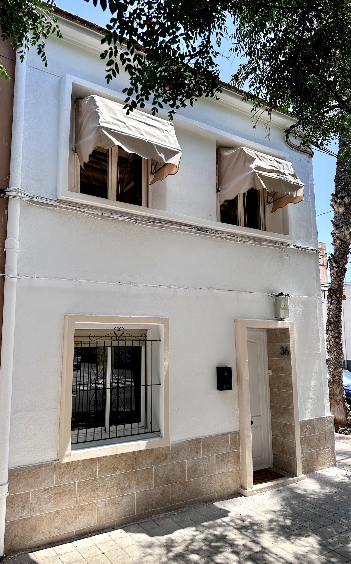 The exterior of a two-story villa features a clean white façade with two windows adorned with awnings. A small entrance door is visible, complemented by a stone base. A decorative wrought iron grille offers additional security for the window below.