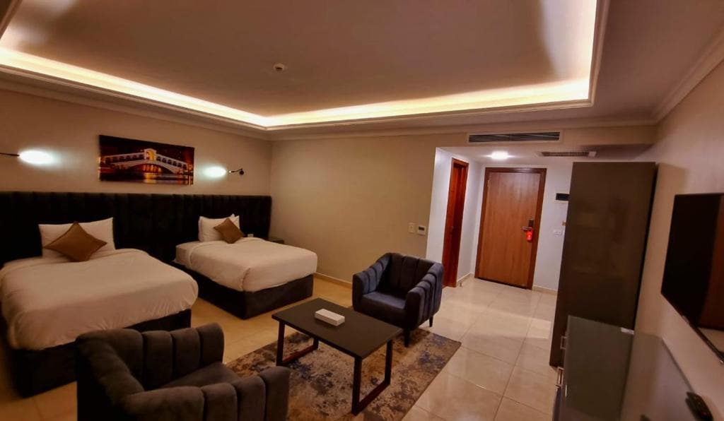 A modern room is presented with two spacious beds, complemented by a sleek dark headboard. A coffee table is positioned in front of comfortable armchairs. Soft lighting highlights the contemporary decor, with a wall-mounted television and a door leading outside visible in the background.