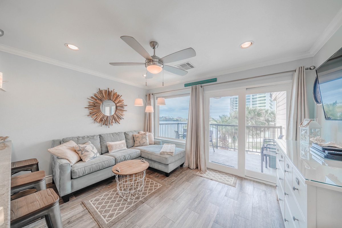 A comfortable living area features a light gray sectional sofa adorned with pillows. A modern coffee table sits in front of the sofa. Large sliding glass doors open to a balcony, allowing natural light to fill the space and offering views of the outdoors.