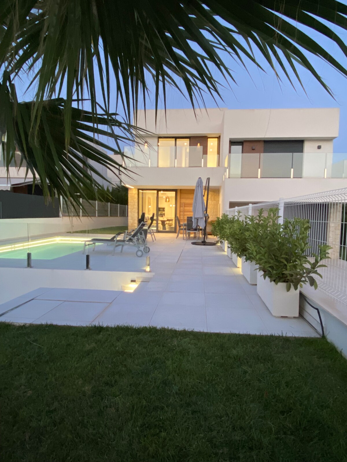 The exterior of the villa is framed by palm leaves, highlighting a well-maintained garden area. A private pool is visible alongside lounge chairs. The terrace features modern outdoor furniture, while large sliding doors suggest an open plan living space illuminated by evening light.