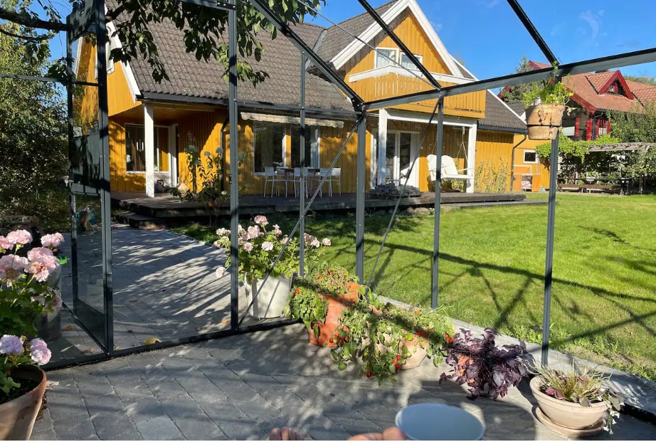 A greenhouse is viewed from the outside, showcasing a paved area with vibrant potted flowers. The yellow house and wooden deck are visible in the background, along with outdoor dining furniture. A spacious lawn is seen, inviting outdoor activities.