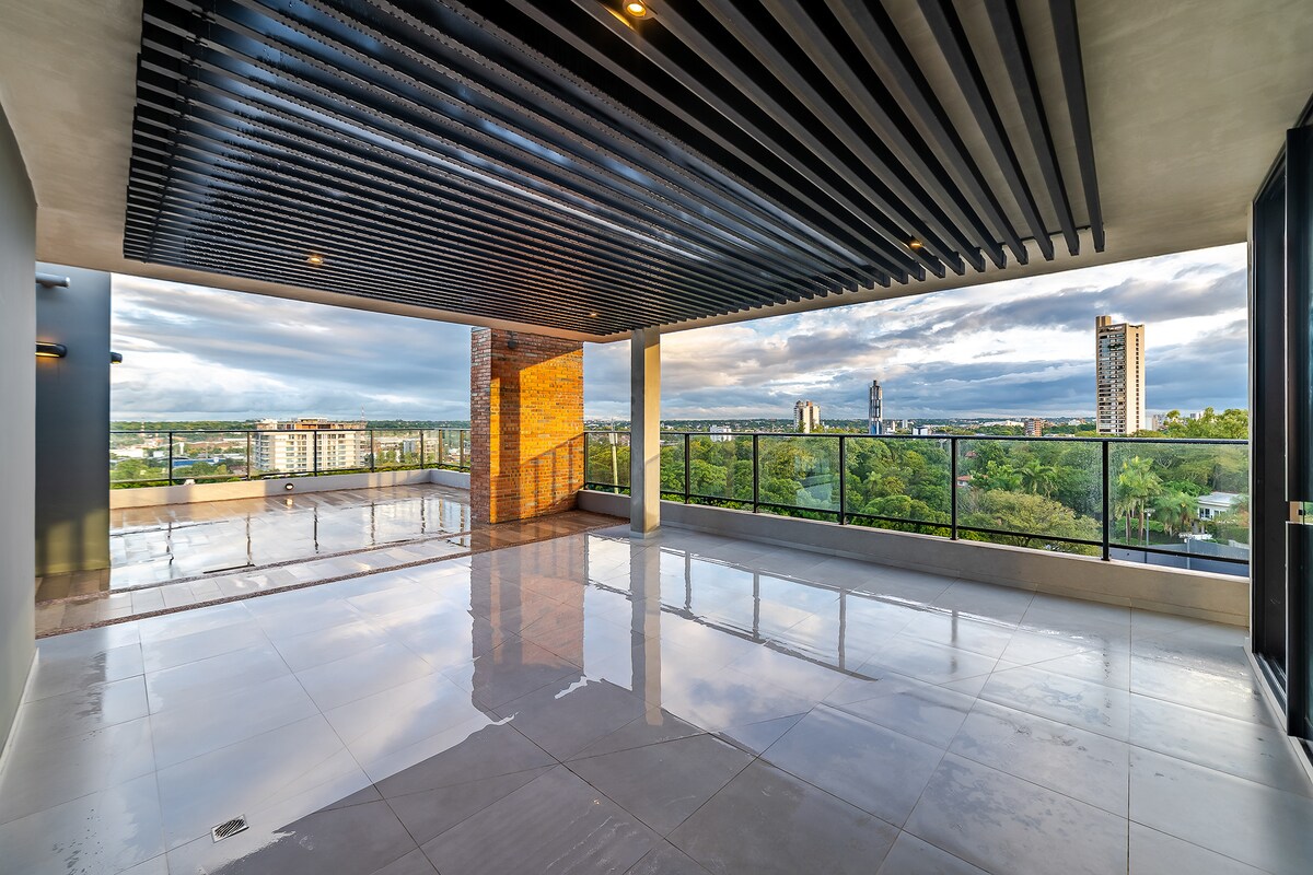An expansive terrace features polished floors and high ceilings, providing an open space with views of the surrounding greenery and cityscape. Large glass doors allow access to the outdoors, while the structural elements showcase a modern design.