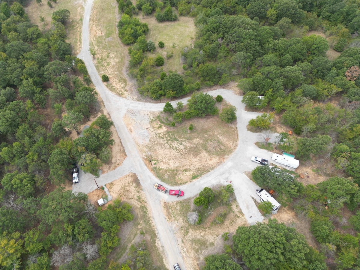 An aerial view shows a network of gravel roads intersecting within a wooded area. Several RVs are parked along the paths, surrounded by green trees and open spaces. The ground is a mix of grass and dirt, suggesting a natural, rustic environment.