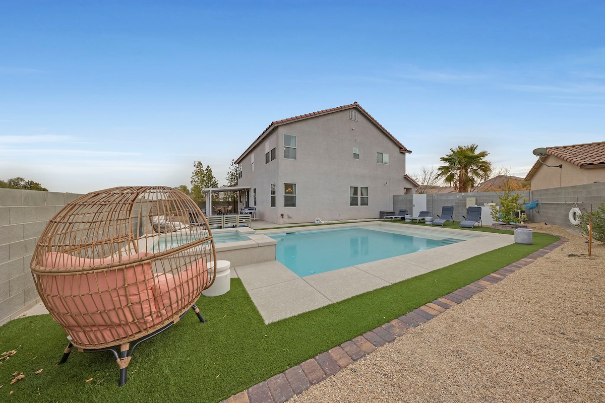 A serene outdoor area features a private pool surrounded by landscaped grass and gravel. A unique wicker seating pod is positioned nearby, creating a comfortable spot for relaxation. The spacious home stands in the background with clear skies overhead, enhancing the tranquil atmosphere.