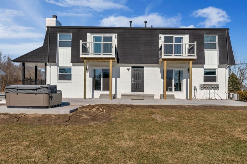 The exterior of a two-story white brick building is shown, featuring a dark roof and wooden balconies. A hot tub is situated on the patio, with expansive lawn space and a clear blue sky above, creating a tranquil outdoor environment.