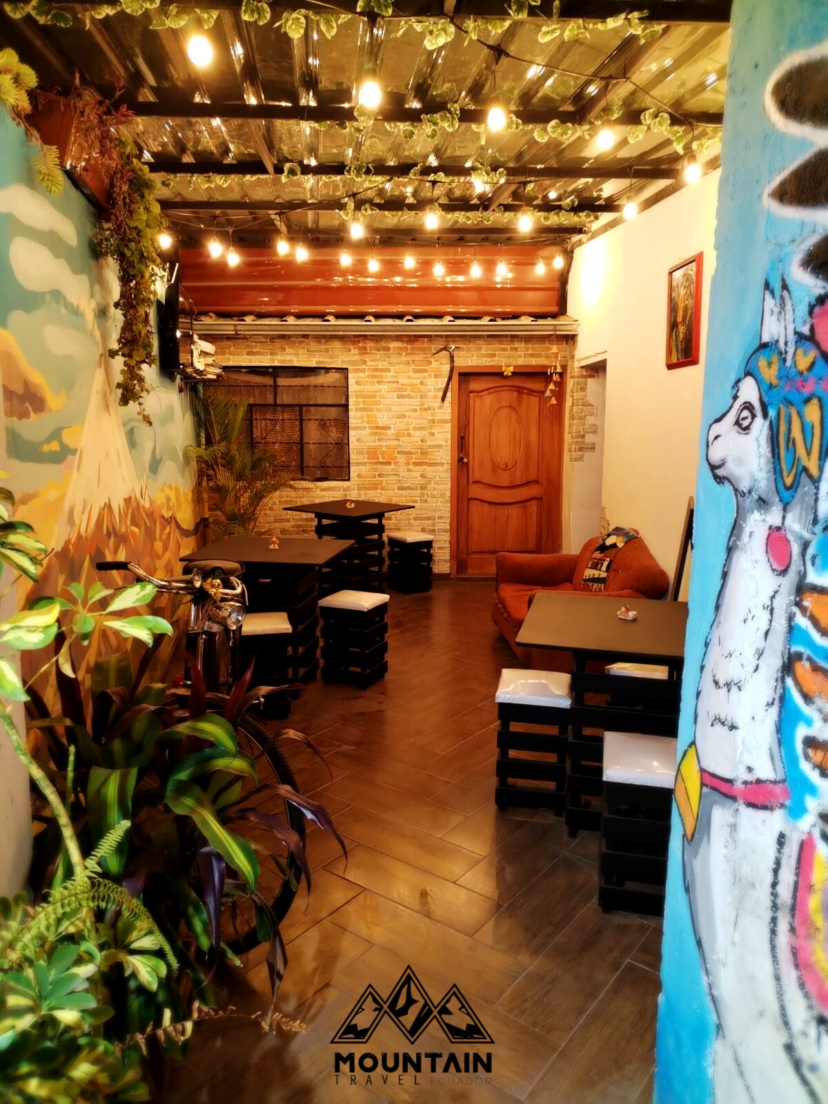A colorful corridor is complemented by warm string lights, enhancing visibility. The walls display vibrant murals, including an artistic depiction of a llama. Various seating arrangements are present, utilizing unique materials like wooden crates, while decorative plants add a touch of nature.