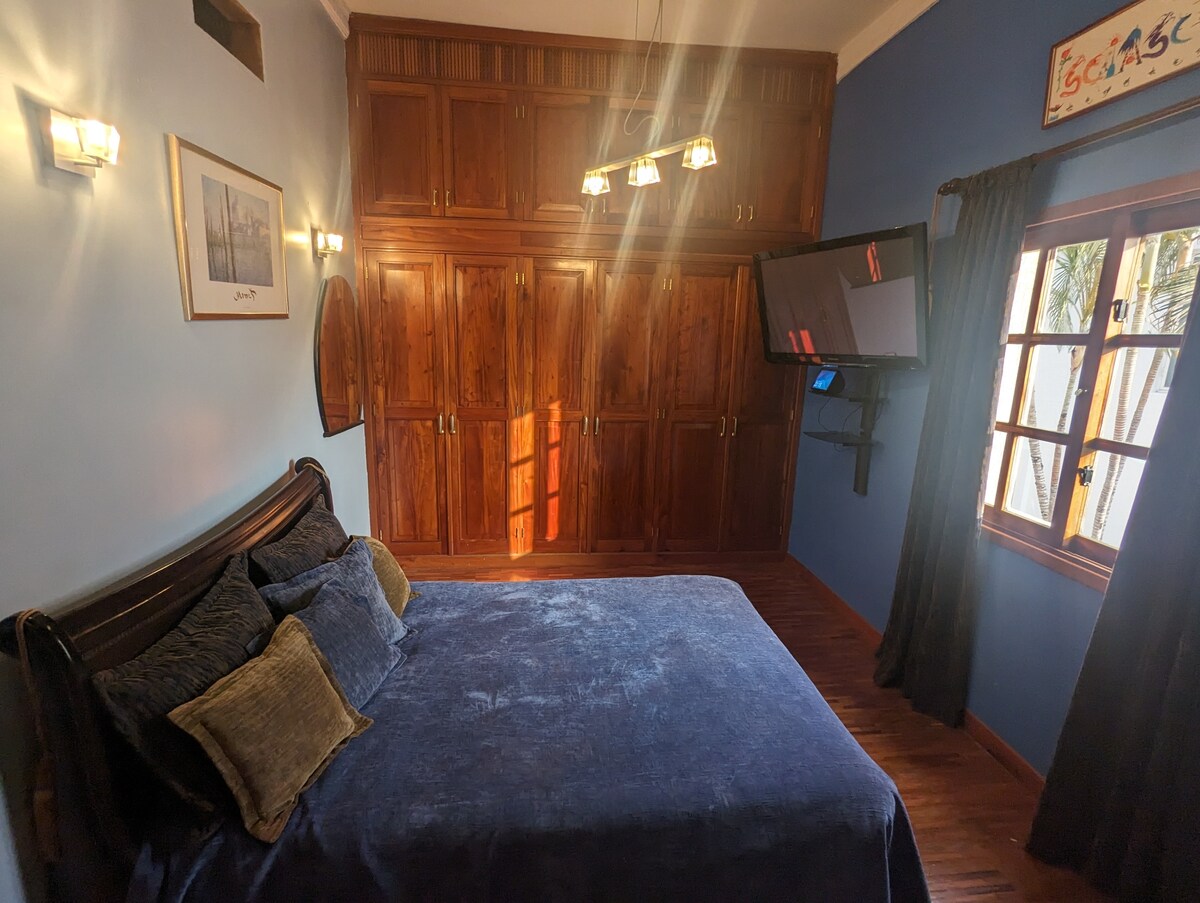A cozy bedroom features a spacious bed with soft blue bedding. Dark wood cabinetry lines one wall, offering ample storage. Natural light filters in through a window with decorative wooden frames, while a wall-mounted TV provides entertainment options.
