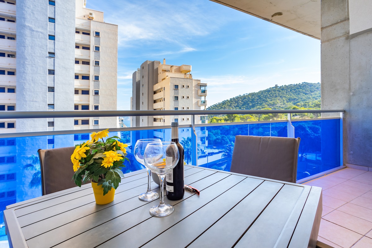 A sunny balcony features a modern table set with two glasses, a bottle of wine, and a yellow flowering plant. The view includes nearby buildings and a green hillside under a clear blue sky.