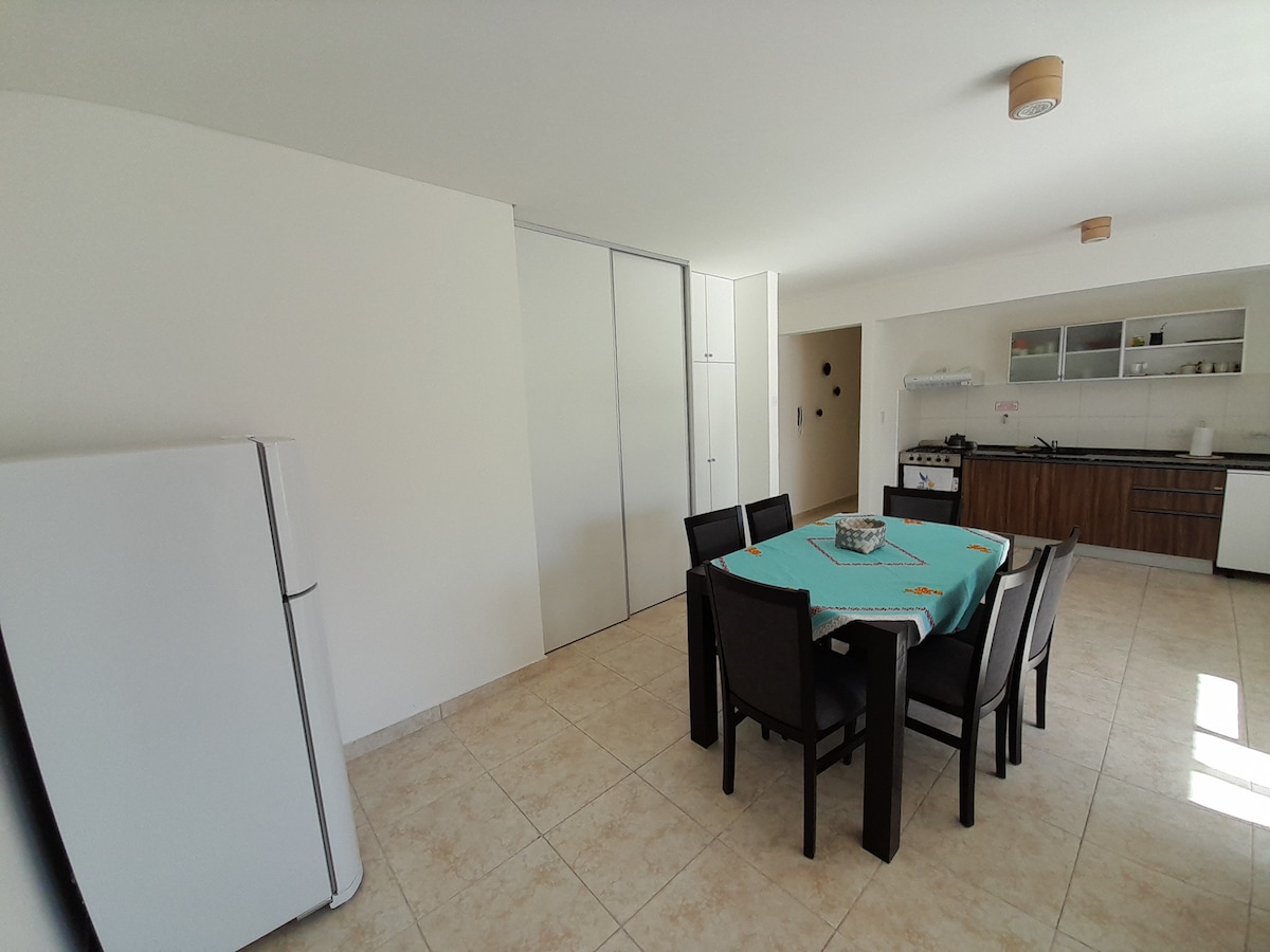 A well-arranged dining area features a rectangular table surrounded by six chairs. A modern refrigerator is visible, alongside a kitchen area equipped with cabinets and cooking appliances. Light-colored tiles cover the floor, contributing to a spacious and bright ambiance.