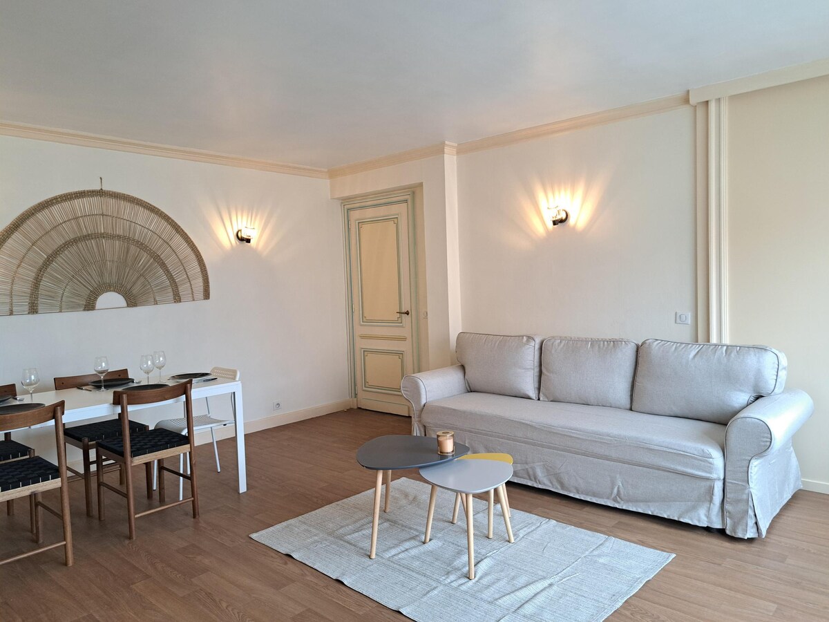 A light-filled living area features a comfortable sofa and a set of wooden dining chairs around a simple table. Soft lighting emanates from wall sconces, illuminating the neutral walls. A small coffee table with decorative elements sits on a light rug, enhancing the spacious feel.