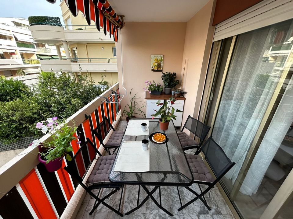 A balcony space features a glass-topped table surrounded by six black chairs. Potted plants add greenery to the area, and bright orange and yellow stripes adorn the awning above. Natural light enhances the tranquil setting, with a view of neighboring greenery.