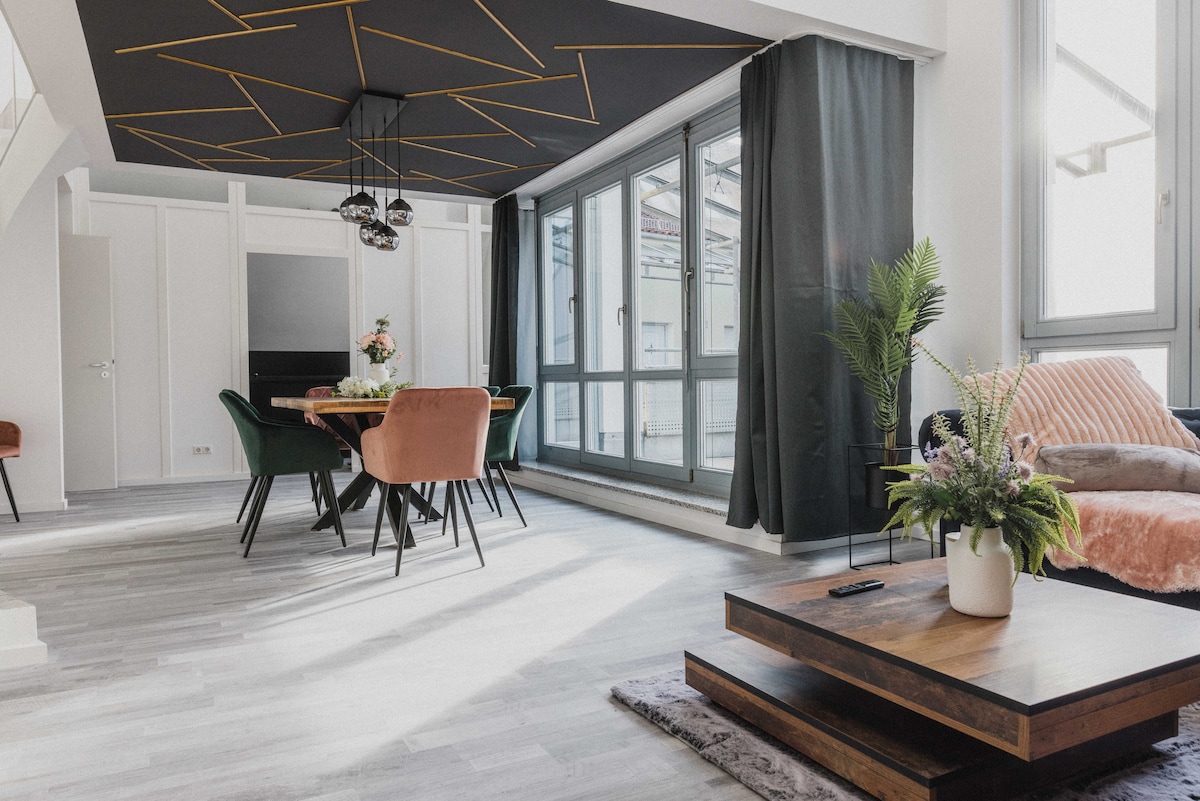 A spacious living area is characterized by large windows allowing ample natural light. A modern dining table with green chairs is complemented by stylish pendant lights. A cozy seating area features a plush sofa, enhancing comfort, while decorative plants add a touch of greenery.