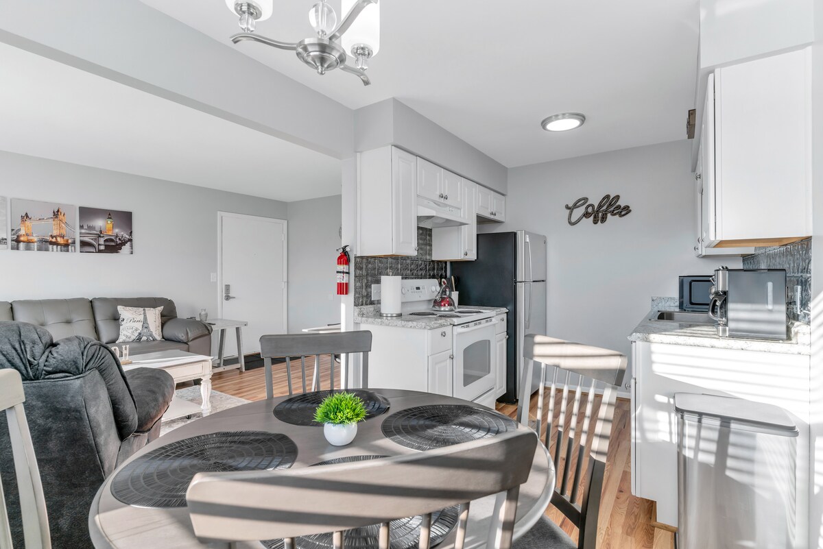A well-lit, modern kitchen and dining area contains white cabinetry and stainless steel appliances. A round dining table with four chairs is positioned beside the kitchen, enhancing the open layout. The living space features a comfortable sofa, with a wall adorned by stylish artwork.
