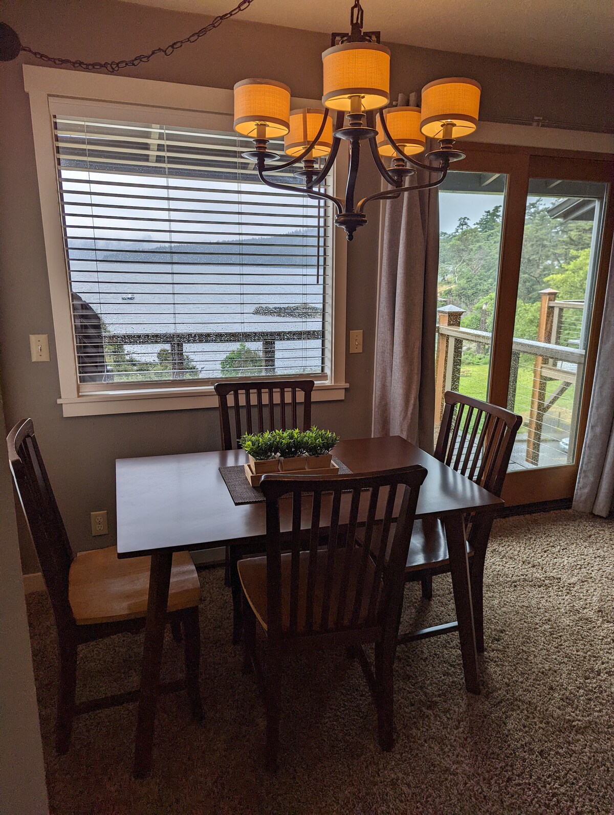 A dining area features a wooden table surrounded by four matching chairs. Natural light filters through the large window adorned with blinds, revealing a view of the water and greenery outside. A simple plant centerpiece enhances the inviting ambiance.