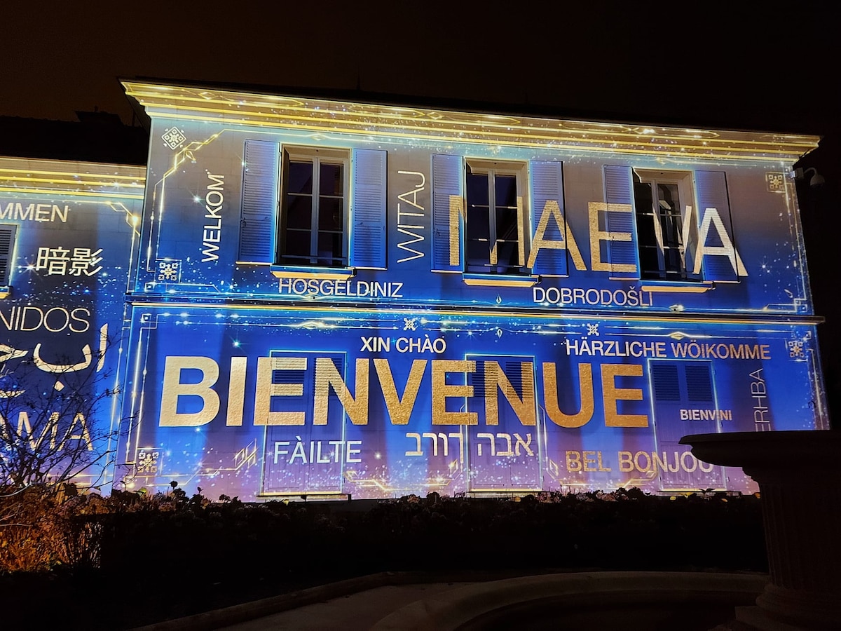 The exterior of a building is illuminated at night, showcasing vibrant projections of the word 'Welcome' in multiple languages. The light display highlights the architecture and creates a festive atmosphere, inviting visitors to engage with the space.
