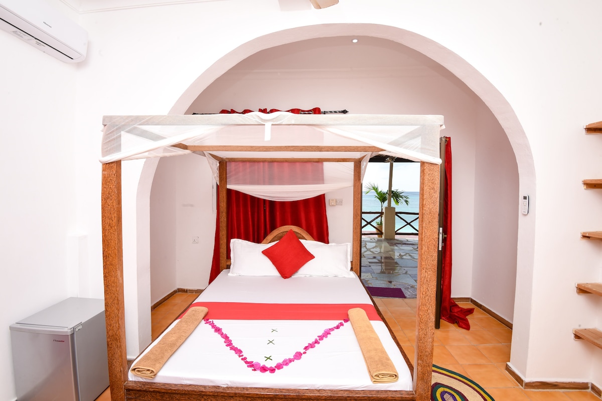 A spacious bed with a decorative canopy is positioned in the room, accompanied by a bright red accent pillow. The walls feature light tones, while a small fridge can be seen to one side. Large windows allow natural light to enhance the ambiance.
