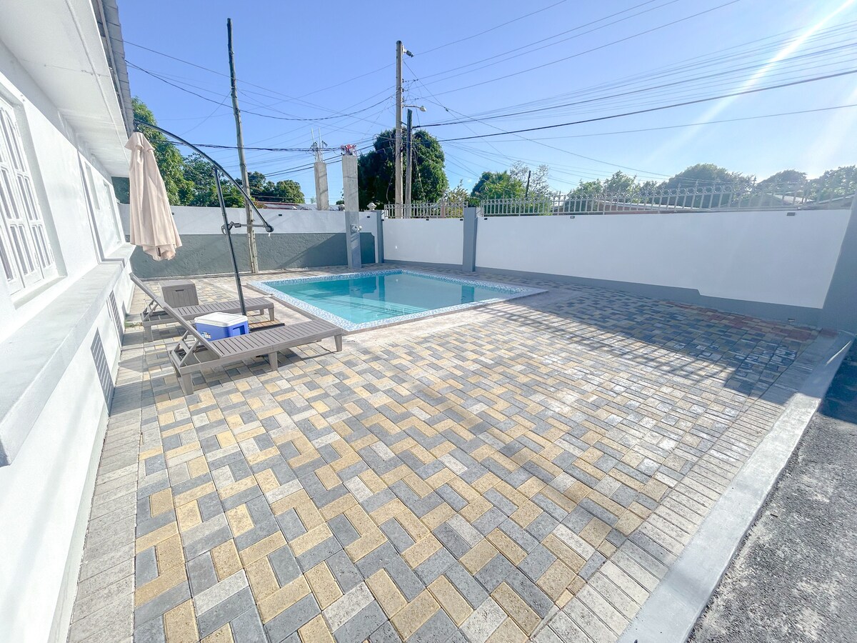 A sparkling pool is bordered by a well-maintained patio featuring a patterned paving design. Lounge chairs are positioned on one side, inviting relaxation in a sunlit outdoor space. The surrounding wall provides privacy, and trees are visible in the background against a clear blue sky.