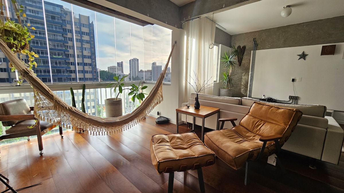 A living area is furnished with a comfortable tan armchair and a cozy lounge chair. A hanging hammock is positioned near large windows, allowing natural light to fill the space. Potted plants add greenery, complementing a modern aesthetic with minimalist decor.