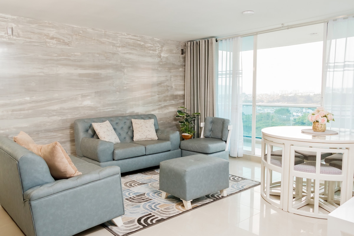 A modern living area is arranged with a comfortable sofa and armchair, set against a textured wall. A round dining table is positioned nearby, complemented by pastel-colored chairs. Large glass doors allow natural light and provide a view of the cityscape beyond.