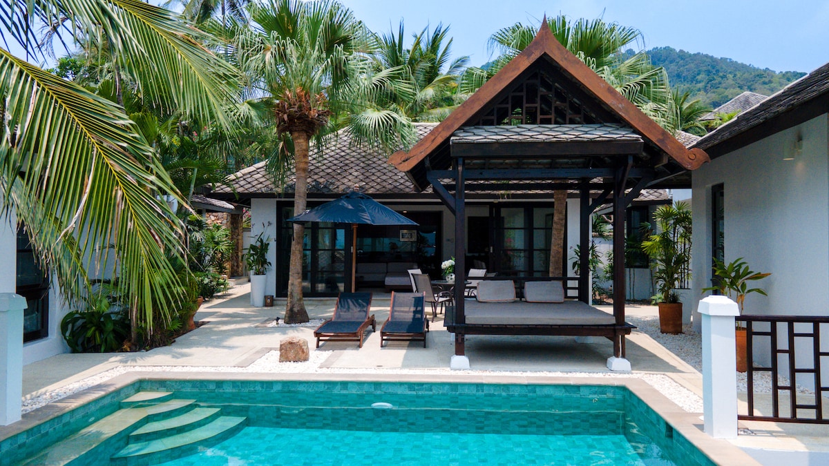 An outdoor area features a private infinity pool surrounded by palm trees. Lounge chairs are positioned next to the pool, while a shaded seating gazebo is present. A pathway leads to the entrance of the villa, showcasing a blend of tropical flora and modern design.
