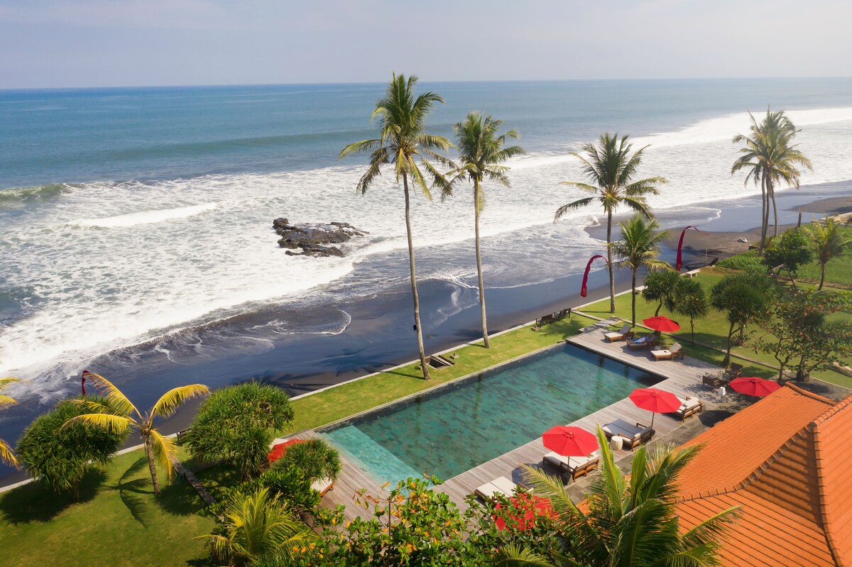 An aerial view captures a serene infinity pool bordered by palm trees and vibrant red umbrellas. The expansive black sand beach and the gentle waves of the ocean can be seen in the background, providing a tranquil setting against the clear blue sky.
