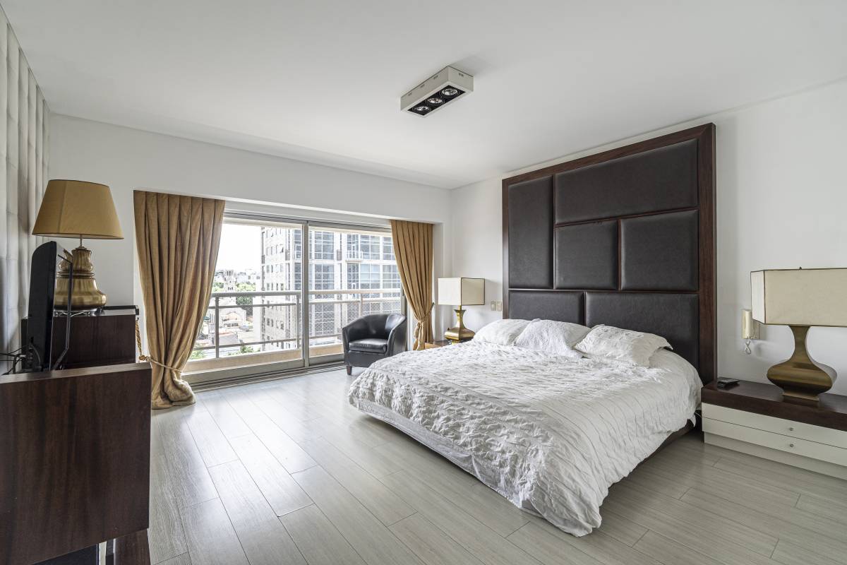 A spacious master bedroom is featured, showcasing a large king-size bed with a textured headboard. Natural light floods the room through a floor-to-ceiling window, framed by elegant curtains. Nightstands with lamps provide additional lighting, and a cozy armchair invites relaxation.
