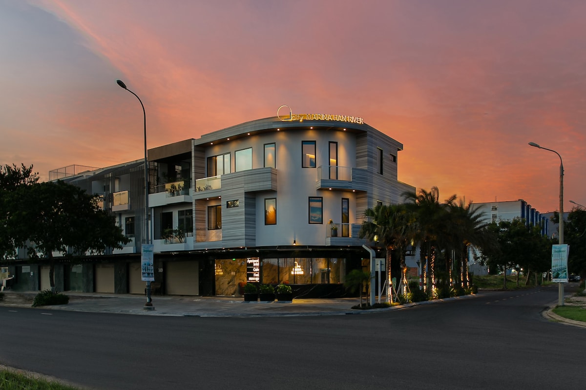 A modern villa is showcased at sunset, featuring a sleek exterior with large windows that reflect the vibrant colors of the sky. Lush landscaping surrounds the building, and warm lighting emanates from the interior, suggesting a welcoming atmosphere.