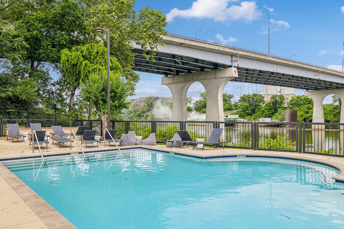 A serene pool area is showcased with clear blue water and inviting lounge chairs positioned around its perimeter. Lush greenery borders the space, and a bridge is visible in the background, connecting the vibrant atmosphere of the surrounding area.
