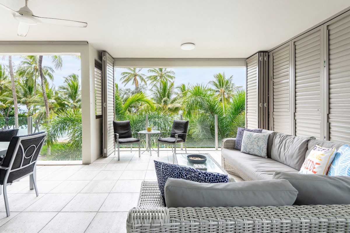 A spacious balcony is furnished with a wicker sofa and two chairs around a small table. Lush tropical greenery is visible through large, sliding glass doors, enhancing the connection between the indoor and outdoor spaces.