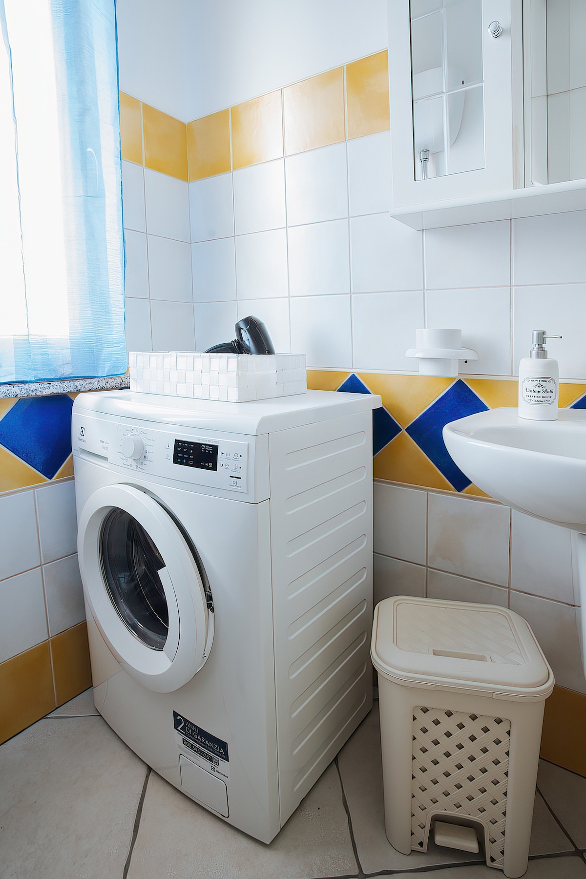 A laundry area is visible, featuring a front-loading washing machine and a small waste bin. Light filters in through a window adorned with blue curtains, illuminating the tiled walls decorated with yellow and blue patterns. A sink is positioned nearby, enhancing functionality.
