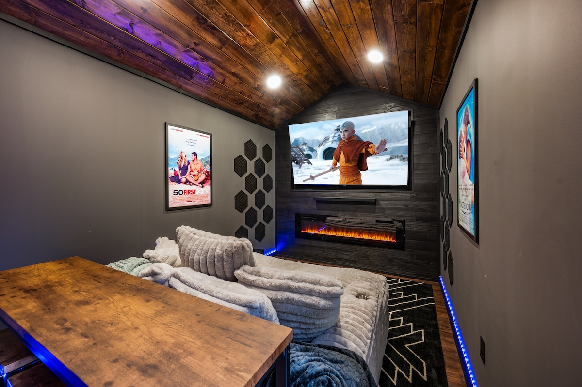 A cozy movie theater room is shown, featuring a large wall-mounted screen displaying a film. A wooden table is positioned in front of a plush sectional sofa, adorned with soft throw blankets. The walls are decorated with movie posters, and LED lighting accents the room's edges.