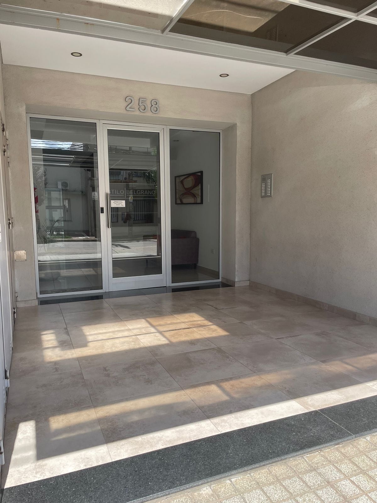 The entrance area features sleek glass doors that allow natural light to enter. A tiled floor is seen in neutral tones, complementing the minimalist design. A small seating area is visible in the background, enhancing the welcoming atmosphere of the space.