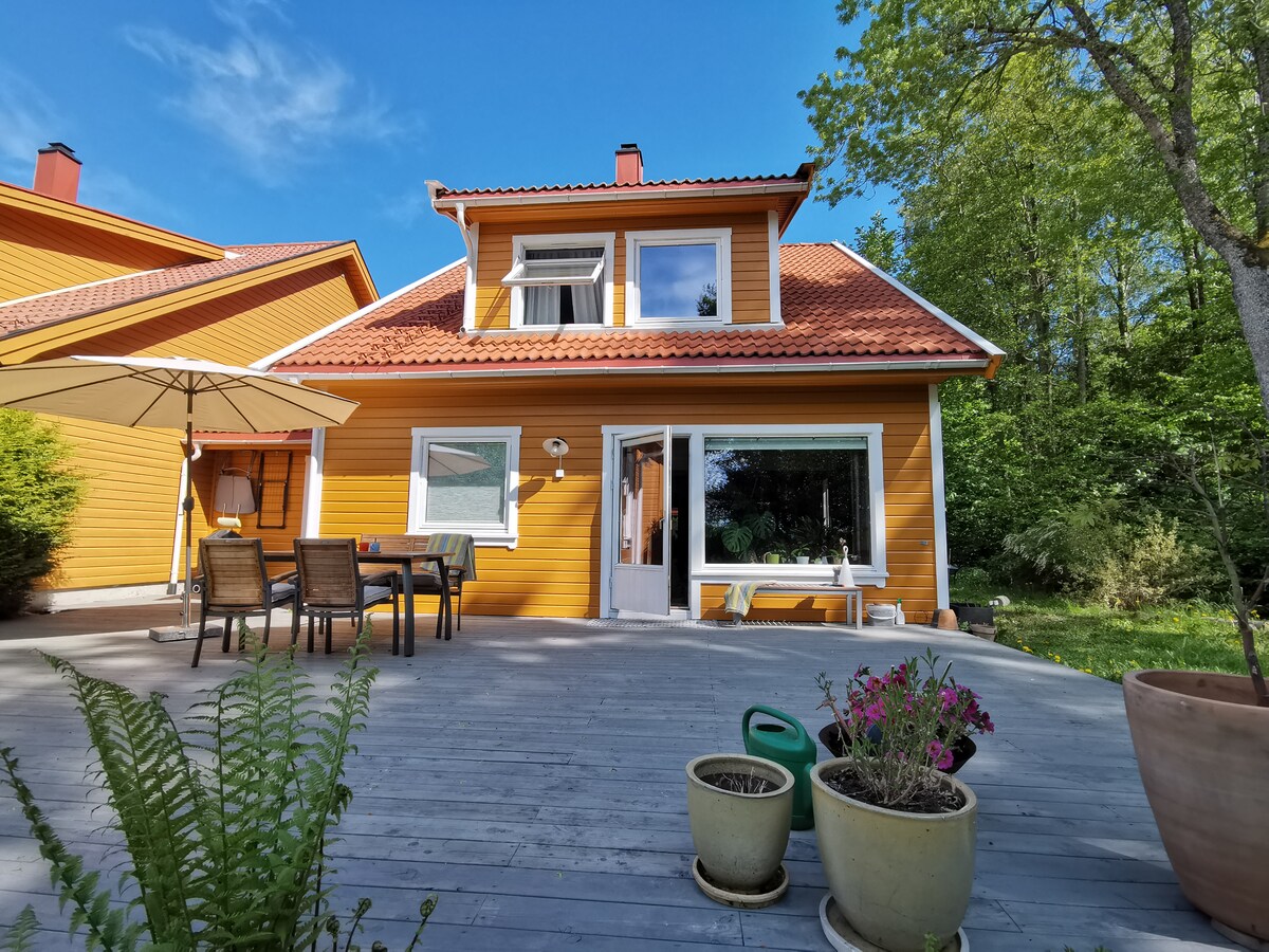 A vibrant yellow house is framed by lush greenery, featuring a large patio space with outdoor furniture and a parasol. Potted plants add a decorative touch to the area, and a doorway offers a glimpse into the inviting interior.