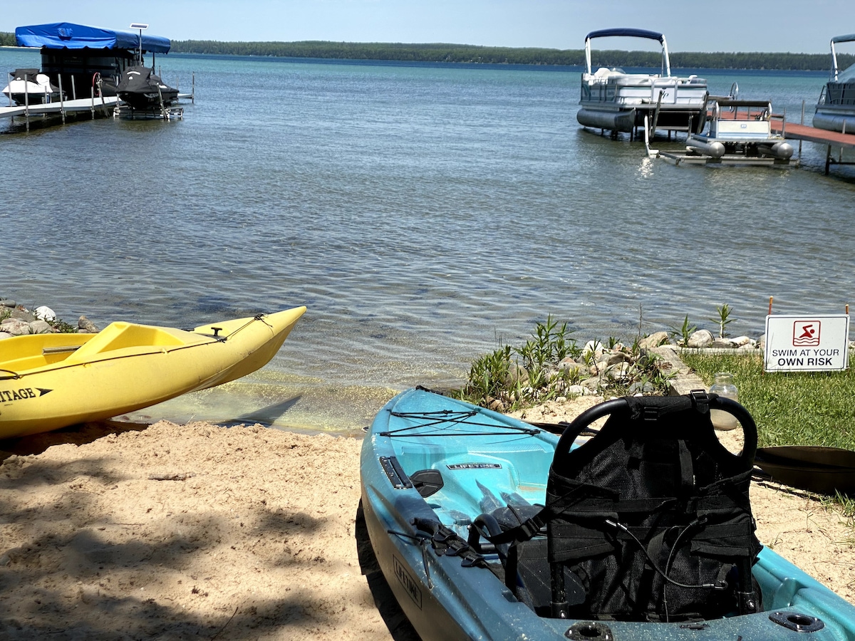 Two colorful kayaks are positioned on a sandy beach, with a calm lake visible in the background. The water reflects blue skies, while a dock is seen in the distance. A warning sign indicates swimming at one’s own risk.