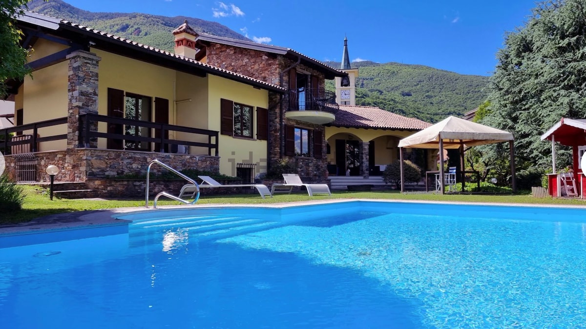 The image features a well-maintained villa surrounded by lush greenery. A sparkling blue pool reflects the sky, with lounge chairs positioned nearby for relaxation. A shaded gazebo offers additional seating, creating a tranquil outdoor space complementing the inviting architecture of the house.