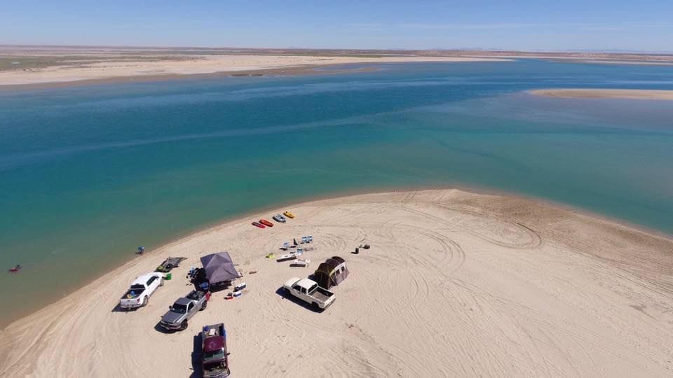 A panoramic view of a sandy beach bordered by shallow turquoise waters. Several vehicles are parked nearby, along with tents and kayaks, indicating a place for relaxation and outdoor activities. The surrounding landscape includes expansive dunes and distant shorelines.